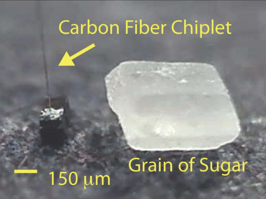 A quartz-like rock, which is really a zoomed-in grain of sugar, sits next to a tiny black chiplet with a wire extending from it. The scale of the chiplet is 150 micrometers.